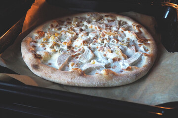 Pizza with pineapple and chicken pieces on a baking sheet on parchment in the oven. Hawaiian pizza on a black background. Cooking at home, the theme of recipes and nutrition