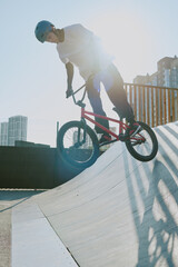 Teenager riding BMX bike on skatepark ramp performing trick wearing helmet outdoors with city...