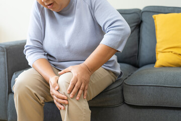 Body arthritis muscles stiff problem, aged woman suffering on leg, pain knee from disease chronic, hand massage rubbing joint injury, mature hurt or sore sitting on sofa at home. Health care people.