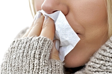 Close-up of a blonde woman in a knitted gray sweater blowing her nose with a white tissue against a bright background. Concept of flu, cold, or seasonal allergy sickness