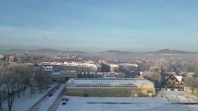 Stadion sportowy zimą widziany z drona. Pokryte śniegiem trybuny i boisko tworzą malowniczy, spokojny krajobraz. Ujęcie z lotu ptaka ukazuje symetrię obiektu i kontrast między bielą śniegu a otoczenie