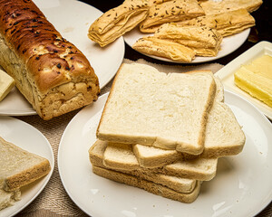 A close-up image of freshly baked bread slices and loaves arranged on white plates with butter and crispy puff pastry snacks. Captured in warm light to highlight texture and freshness.