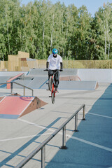 Teenager performing trick on BMX bike at outdoor skatepark, wearing helmet and gloves, balancing on rear wheel near metal rail, trees in background