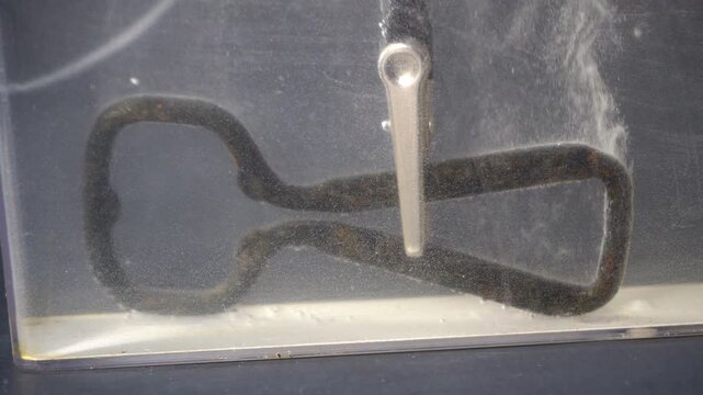 Rusty bottle opener releases bubbles during electrolytic cleaning. Patches of rust are replaced with new grey metal.