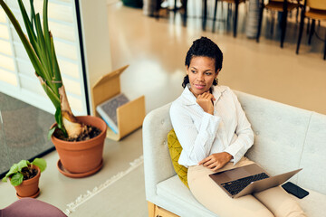 African-American Woman Working on Laptop in Modern Office Lounge, Thinking, Seated on Couch in Cozy...