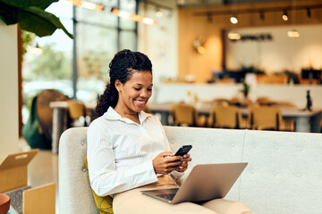 African-American Woman Working on Laptop While Smiling on Sofa in Modern Café