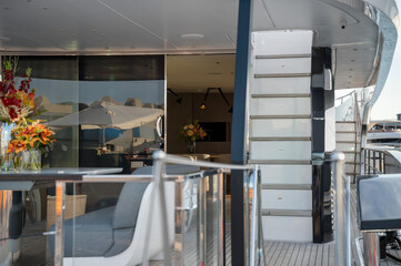 Modern yacht exterior with glass railings, cozy gray seating, floral arrangements, and a sleek staircase leading to the upper deck at Limassol Marina.
