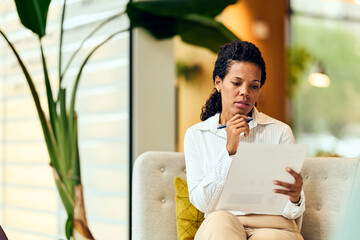 African-American Woman Reviewing Documents in a Modern Office Lounge, Thinking Deeply on Paper