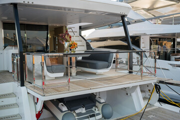 Modern yacht deck with comfortable grey cushioned seating, glass railings, and floral arrangements in vases. Teak flooring and nearby docked boats visible in a peaceful marina.
