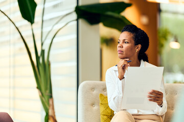 African-American Woman Thinking With Documents In Hand In Modern Office Lounge