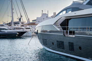 Luxury yachts moored closely in a tranquil marina at sunset. The dark hulls reflect the calm blue water, with sleek lines and spacious decks visible at Limassol Marina.