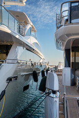 Two luxury yachts docked side by side at a marina, with polished hulls reflecting the calm blue water. Teak decks and protective black fenders line the sides at Limassol Marina.