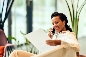 African-American Woman Smiling While On Phone And Reviewing Paperwork In Modern Office Setting
