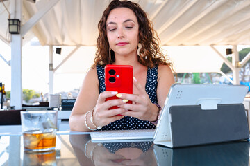 Elegant adult woman with curly hair sitting at glass table outdoors using red smartphone with drink...