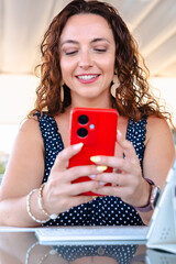 Elegant adult woman with curly hair sitting at glass table outdoors using red smartphone with drink...