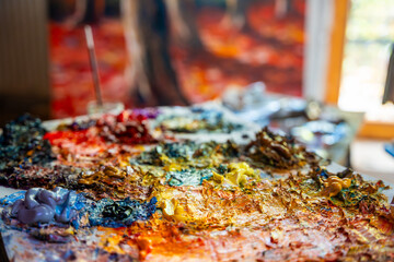 Close-up of a messy painter palette with thick dabs of oil paint in various bright colors. In the background, a blurry painting is visible, evoking creativity and inspiration.