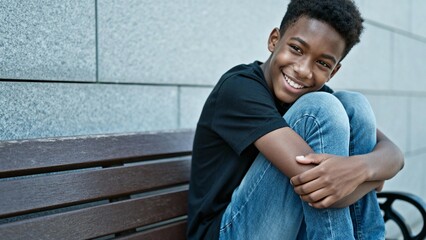 Smiling black teenager sitting on wooden bench with knees hugged  