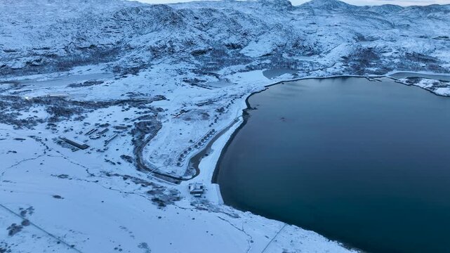 Aerial view of a remote arctic landscape with a cluster of buildings and a row of small domes along the shore, surrounded by vast snow-covered mountains and a large body of water