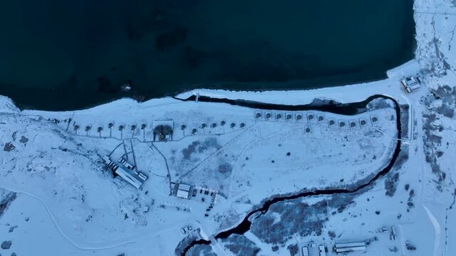 Top down aerial view of a remote arctic camp or settlement with a row of geodesic domes, various buildings, and a dark, winding river cutting through the vast snow-covered landscape