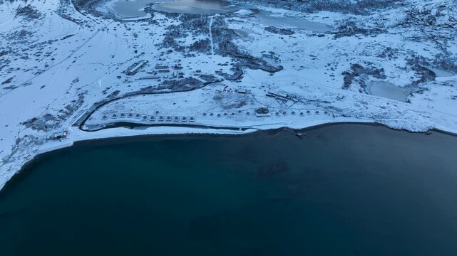 Aerial view of a remote arctic landscape with a row of small domes or tents along the coastline, snow-covered hills, and frozen lakes inland; a small part of a computer screen interface is visible on 