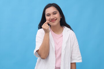Fototapeta premium Close-up portrait of young Asian woman wearing casual clothes, making Korean love sign with fingers, smiling looking at camera, standing isolated on blue background.