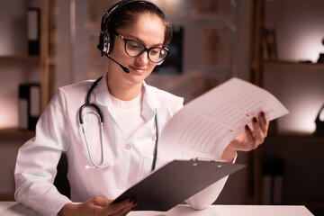 female doctor in white coat with headset and clipboard consulting patient via video call making online healthcare conference, reading list of medicine in hospital office
