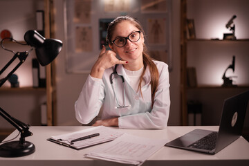 female doctor in glasses and white coat working on night shift, sitting at desk showing phone sign...