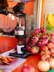 Hands wearing gloves press fresh pomegranates in a juicer at a vibrant market stand filled with colorful fruits, including dragon fruit and carrots