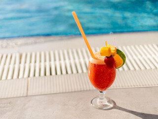 A colorful tropical cocktail is placed on a stone surface next to a pool. The drink features vibrant garnishes and a straw, perfect for enjoying under the sun. Copy space.