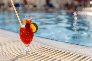 A refreshing tropical cocktail sits on the edge of a pool, adorned with fresh fruit and a colorful umbrella. Sunlight reflects on the water, creating a vibrant atmosphere