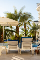 Two white lounge chairs with blue cushions sit beside a sparkling pool surrounded by tall palm trees and clear blue skies. A large umbrella provides shade, creating a serene oasis