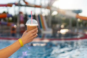 A hand holds a cup of iced drink with a pink straw, set against the backdrop of a vibrant water park. Features colorful water slides and a sparkling pool. All inclusive.
