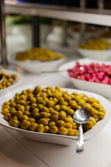 A large bowl filled with green olives sits in the foreground, surrounded by various dishes with colorful pickled vegetables. The setting is a buffet, inviting guests to enjoy the food