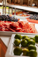 Colorful slices of watermelon and vibrant limes are arranged neatly on white trays at a lively fruit buffet. The setting is bright and inviting, perfect for a summer gathering