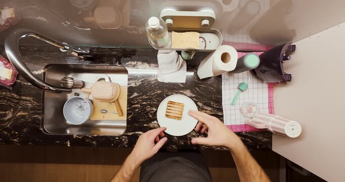 A man prepares a sandwich in his kitchen and accidentally burns his fingers on the hot toast. The scene captures a relatable everyday kitchen mishap from a top-down perspective.