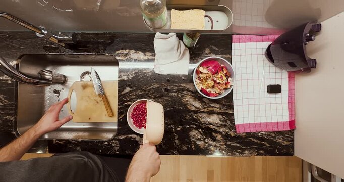 A man pours freshly washed pomegranate seeds from a container into a metal bowl on a kitchen counter. The bright red fruit contrasts beautifully with the clean kitchen setup