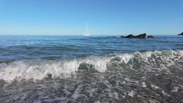 Waves gently lap on the shore of Portnadler Bay in Cornwall, near Looe and Talland. A sail boat is is anchored in the cove. Beautiful sunny day. Shingle beach. 