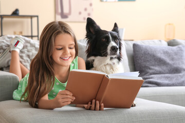 Cute little girl with Border Collie dog reading book on sofa at home