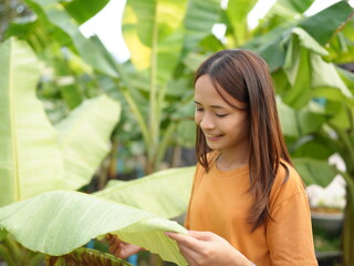 Young woman examining large green leaves in a lush garden, showcasing her interest in nature and plant life with a joyful expression on her face