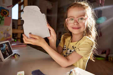 Smiling girl proudly shows her drawing in a sketchbook.
Creative child enjoys art and self-expression at home.
Sunlight highlights happy learning and imagination moment.

