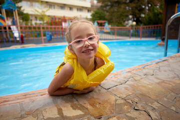 Smiling girl in yellow lifejacket enjoys swimming pool fun.
Happy child with pink glasses leans on pool edge on summer day.
Outdoor leisure moment captures childhood joy, safety, and sunshine.
