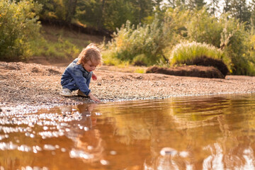 Little child in denim explores riverbank on a sunny day.
Curious toddler touches clear water surrounded by nature.
Peaceful outdoor moment reflects childhood freedom and joy.

