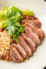 A sliced seared duck breast with fresh herbs, nuts, and greens is presented on a minimalist plate against a dark background, captured from a top-down view
