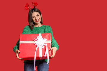 Fototapeta premium Young woman in reindeer horns with Christmas gift on red background