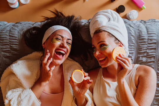 Two happy women lying on bed are applying beauty products and making peace sign - Powered by Adobe