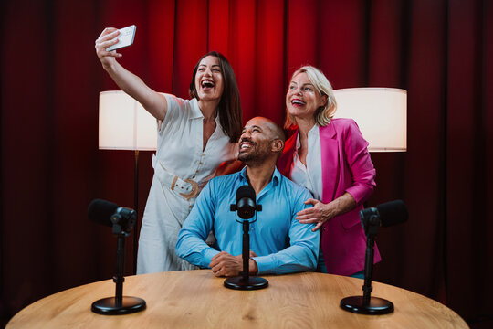 Group of hosts is capturing a live audio broadcast, engaging in a lively, amusing conversation around microphones and a laptop in a professional studio setting