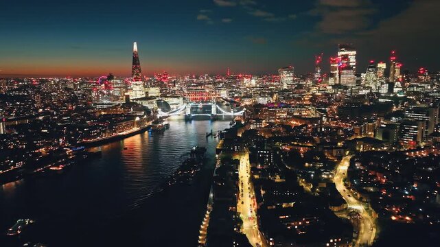 England, London: Breathtaking aerial view of night London city with Tower Bridge, The Shard, and other landmarks in colorful bright lights creating a stunning cityscape skyline under sunset sky