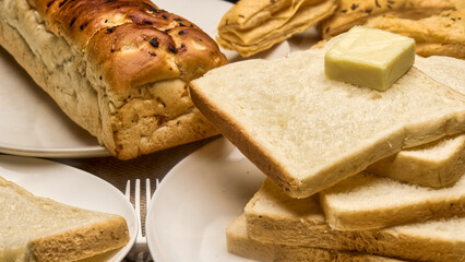 Close-up image showcasing a freshly baked golden brown bread loaf beside soft white bread slices stacked on a plate with a cube of butter on top. Crispy puff pastries in the background