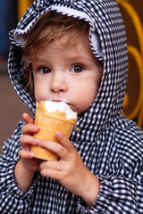 Adorable toddler in a checkered hood enjoys creamy ice cream outdoors. 
Closeup portrait captures sweet childhood innocence and emotion.
Natural light highlights cozy autumn street moment.