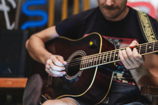 A young Caucasian man with a beard plays an acoustic guitar. He wears a black t-shirt and is focused on his music in a casual setting.
